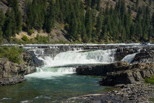 A scenic view of a waterfall cascading over rocky cliffs, surrounded by lush green trees and a clear blue sky.