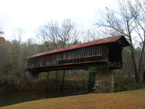 A weathered covered bridge with a rusted roof spans a calm river, surrounded by bare trees and autumn foliage.