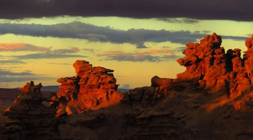 Sunset over unique rock formations, casting warm orange hues against a dramatic sky with clouds.