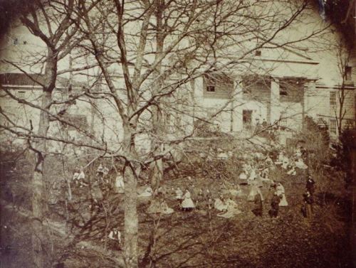 A historic black-and-white photo of a large group of children sitting outdoors near a building, surrounded by trees.