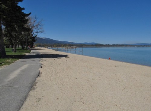 A sandy beach with a clear blue sky, trees lining the path, and mountains in the distance. Calm water reflects the scenery.
