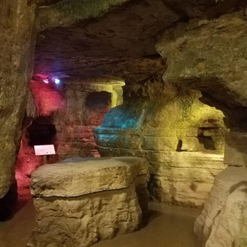 A dimly lit cave interior with colorful lights illuminating stone formations and a sign in the background.