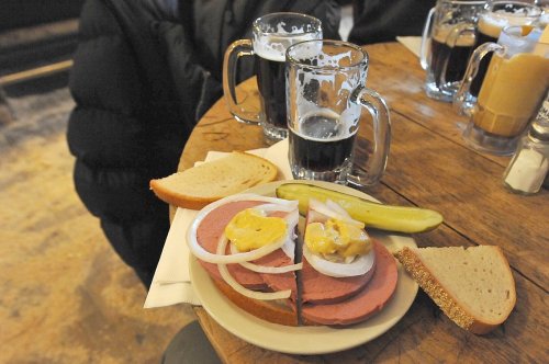 A plate with sliced meat, onions, mustard, and pickles, accompanied by two glasses of dark beer on a wooden table.