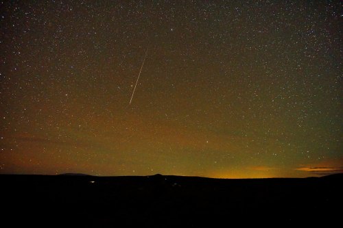 A starry night sky with a bright meteor streaking across, set against a dark landscape.