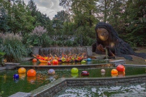 A serene garden scene featuring a colorful glass sculpture in a pond, with a large stone figure in the background.