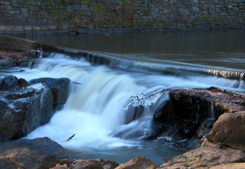 A serene waterfall cascades over rocks, surrounded by a calm river and icy formations along the edges.