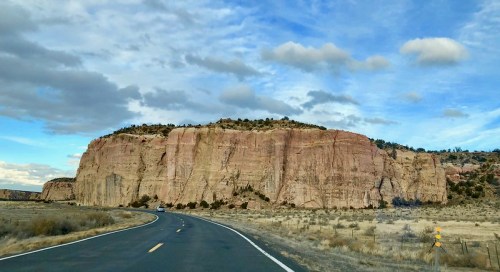 A winding road leads past a large, colorful rock formation under a partly cloudy sky.