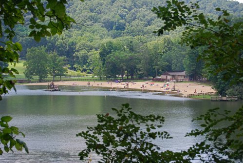 A serene lake view with a sandy beach, surrounded by green trees and people enjoying the sun.