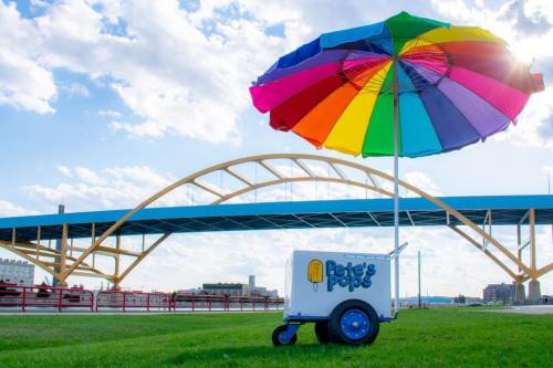 A colorful umbrella shades a popsicle cart in front of a blue bridge and a bright sky.