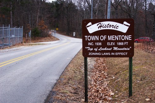 Sign for the historic Town of Mentone, elevation 1868 ft, with a winding road and trees in the background.