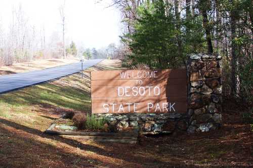 Wooden sign reading "Welcome to Desoto State Park" beside a road, surrounded by trees and natural scenery.