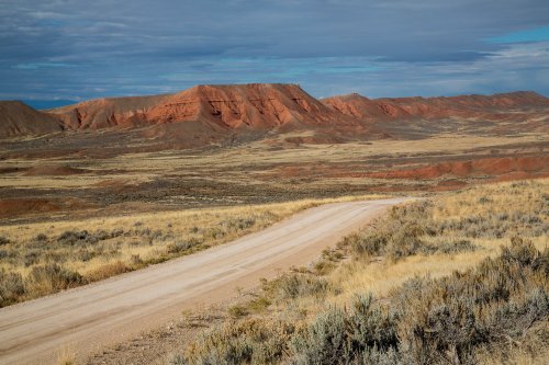 A winding dirt road leads through a vast, arid landscape with red rock formations and sparse vegetation under a cloudy sky.