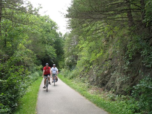 Two cyclists ride on a scenic path surrounded by lush greenery and rocky terrain.