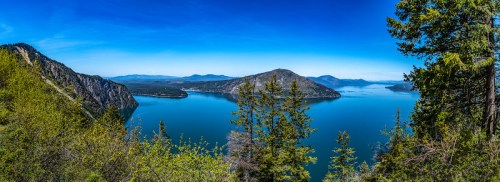 A panoramic view of a serene lake surrounded by mountains and lush green trees under a clear blue sky.