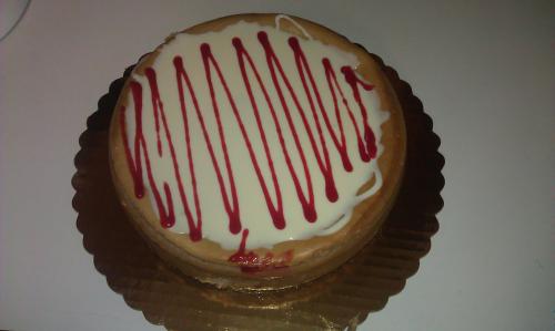 A round cake with a creamy white frosting and red drizzle on top, placed on a gold cake board.
