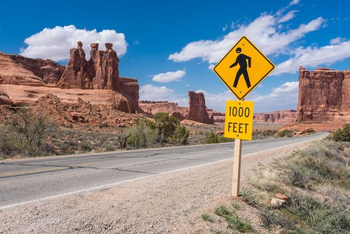 A yellow pedestrian warning sign stands beside a road in a desert landscape with red rock formations and blue skies.