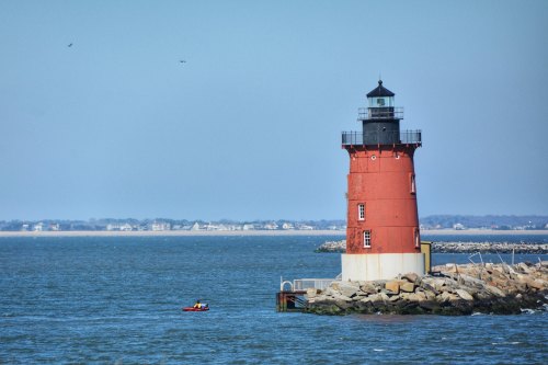 A red and black lighthouse stands on a rocky shore, with a small boat nearby and a clear blue sky in the background.