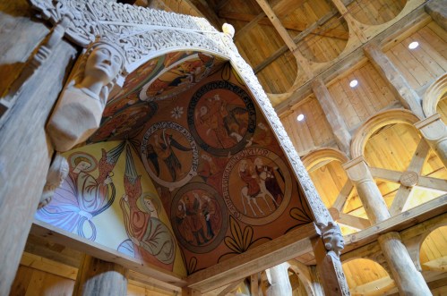 Interior view of a wooden structure featuring intricate carvings and colorful murals on the ceiling.
