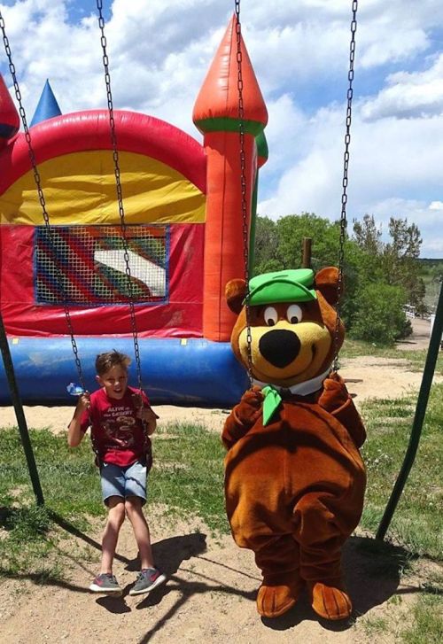 A child swings next to a costumed bear character at a playground with a bouncy castle in the background.