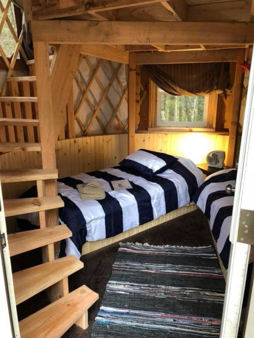 Cozy wooden interior of a yurt with two beds, a window, and a spiral staircase leading to a loft.
