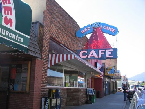 A colorful cafe sign reading "Red Lodge Cafe" on a brick building, with shops and mountains in the background.