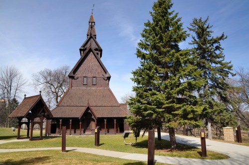 A wooden church with a tall spire, surrounded by trees and a grassy area under a clear blue sky.