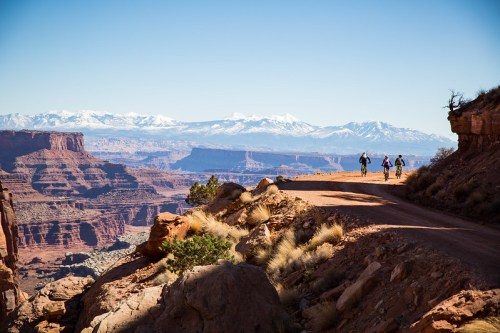 Two cyclists ride along a dirt path with stunning mountain views in the background under a clear blue sky.