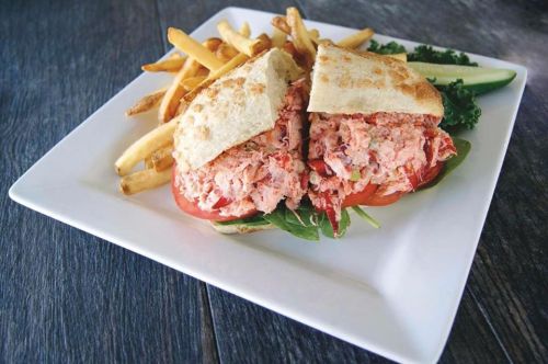 A lobster sandwich with tomato and greens, served with fries and a cucumber slice on a white plate.