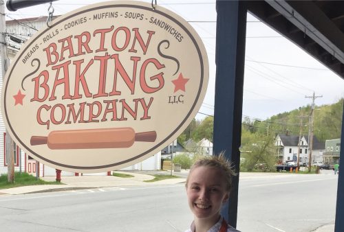 A smiling young chef in an orange apron stands outside the Barton Baking Company, with a sign overhead.