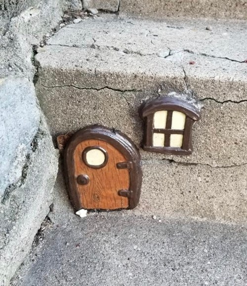 A small, whimsical door and window set into a concrete step, resembling a miniature fairy house.