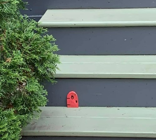 A small red object resembling a face is embedded in a green wooden step, next to a bush.