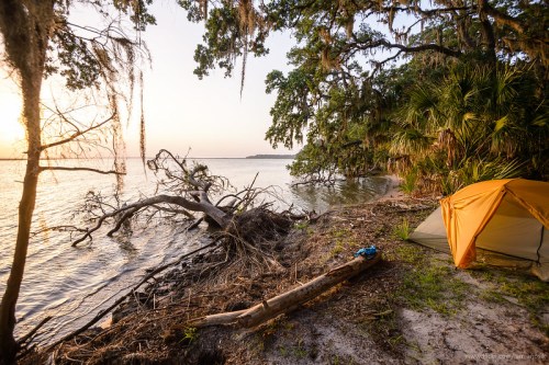 A serene campsite by the water at sunset, featuring a tent, trees, and a calm shoreline.
