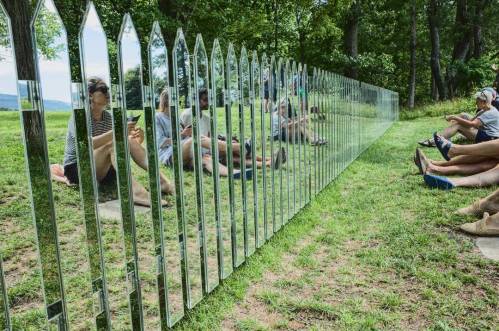 A mirrored fence reflects people sitting on the grass, surrounded by trees and a scenic landscape.