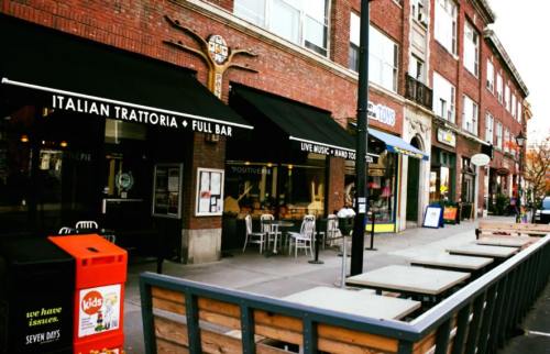 Street view of a vibrant neighborhood featuring an Italian restaurant, outdoor seating, and various shops.