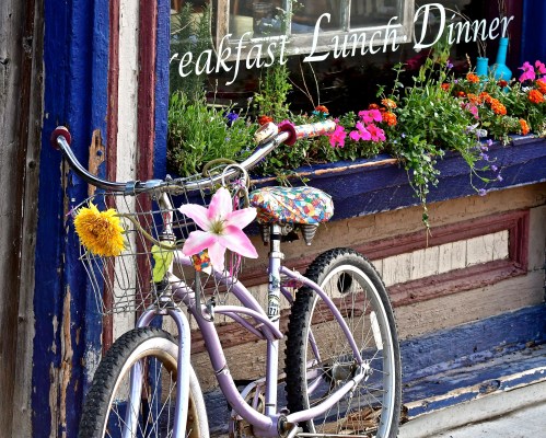 A purple bicycle with flowers in the basket parked outside a restaurant with colorful window flowers.