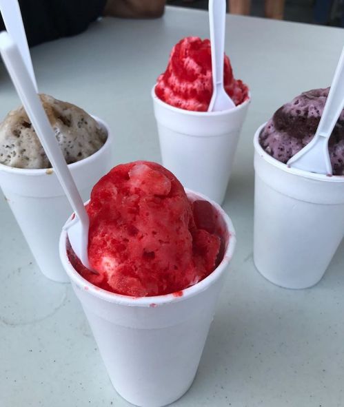 Four cups of colorful shaved ice desserts, with flavors including red, gray, and purple, each with a white spoon.