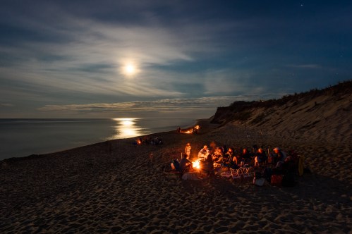 A group gathers around a campfire on a beach at night, illuminated by moonlight reflecting on the water.