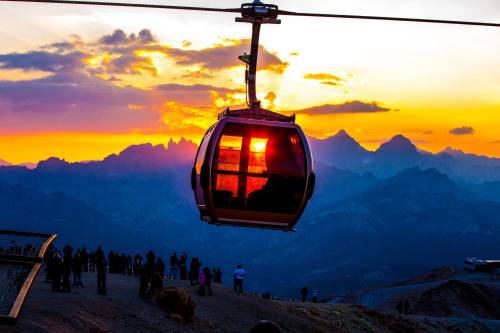 A gondola glides over mountains at sunset, with silhouettes of people watching below.