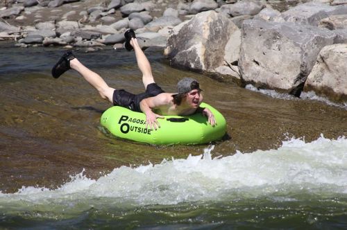 A person in a green inner tube is floating down a river, arms outstretched, with rocks and water splashing around.