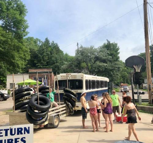 A group of people gather near a bus, preparing for a day of tubing, with inflatable tubes stacked nearby.