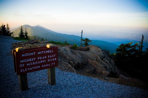 Sign at Mount Mitchell, highest peak east of the Mississippi River, with a scenic view of mountains in the background.