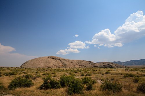 A large rock formation rises above a grassy landscape under a blue sky with scattered clouds.