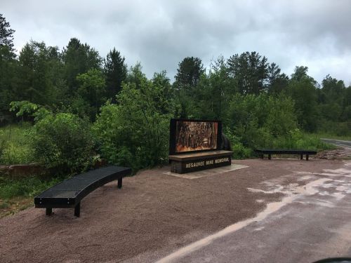 A memorial site with a wooden display and benches, surrounded by lush greenery and overcast skies.