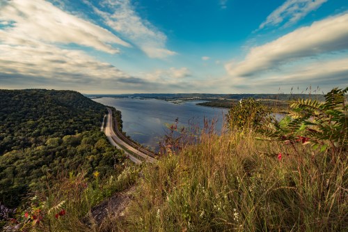 A scenic view of a river winding through lush green hills under a blue sky with scattered clouds.