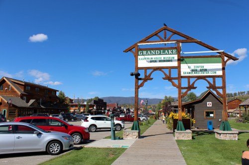 A scenic view of Grand Lake, featuring a welcome sign, shops, and parked cars under a clear blue sky.