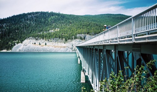 A long bridge extends over a turquoise lake, surrounded by green hills and trees under a blue sky.