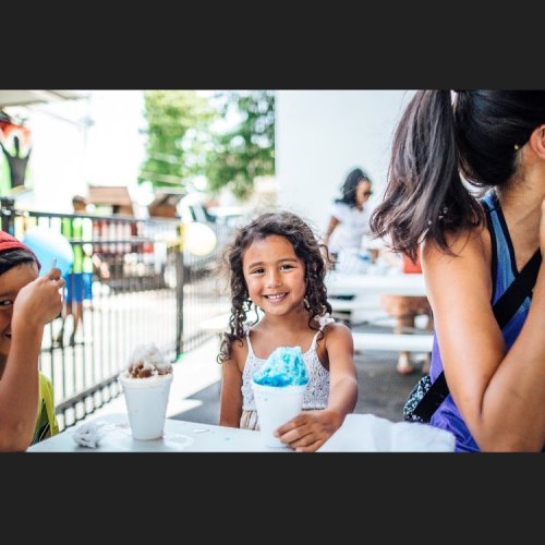 A smiling girl holds a cup of blue snow cone, sitting at a table with others enjoying treats on a sunny day.