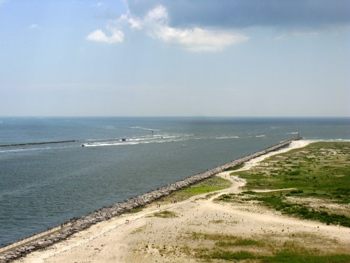 A coastal view featuring a jetty extending into the water, with boats and sandy shorelines under a partly cloudy sky.
