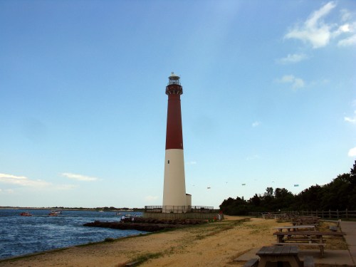 A tall lighthouse stands by the shore, with a sandy beach and calm water under a clear blue sky.