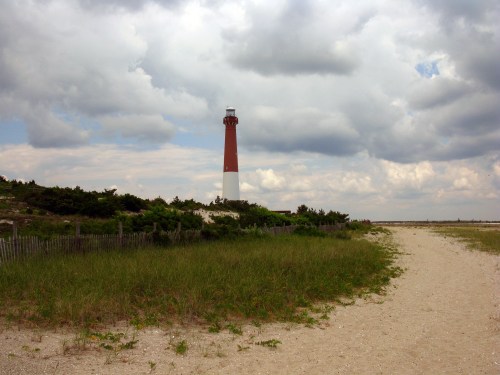 A red and white lighthouse stands tall against a cloudy sky, surrounded by sandy paths and green grass.